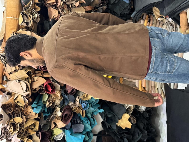 May include: A brown suede jacket with a structured collar and long sleeves, worn over a dark red shirt and blue denim jeans. The jacket features visible seam detailing along the back and shoulders, providing a clean and fitted appearance.