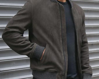 May include: A dark olive green bomber jacket featuring a full-length front zip, a ribbed collar, ribbed cuffs, and a ribbed hem in a charcoal grey tone. The jacket includes two side pockets and is worn over a plain dark-coloured shirt.