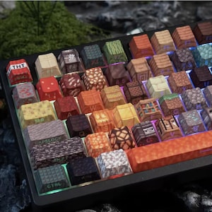 Peut inclure: Un clavier mécanique personnalisé avec des touches conçues pour ressembler à des blocs d'un jeu vidéo populaire. Les touches présentent diverses textures et couleurs, notamment du bois, de la pierre et du TNT. Le clavier a un cadre noir et une barre d'espace orange.