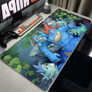 May include: A rectangular desk mat featuring a vibrant illustration of blue and green cartoon creatures in a watery environment. The design includes a large blue character with orange accents, surrounded by smaller, similar creatures. The mat is designed for use with a computer.