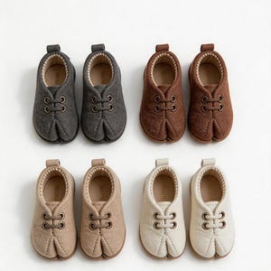 May include: Four pairs of split-toe shoes arranged in two rows. The top row features charcoal gray and brown pairs, while the bottom row includes beige and off-white pairs. Each shoe has a rounded toe, a central seam, and two metal eyelets.