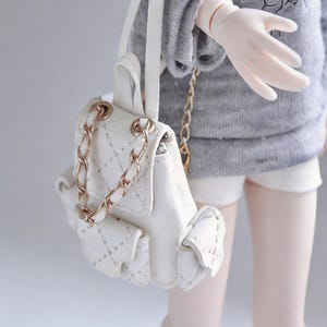 Puede incluir: Una pequeña mochila acolchada blanca con correas y detalles de cadena dorada. La mochila tiene un cierre de solapa y bordes decorativos con volantes. La bolsa se muestra sobre un fondo gris suave.