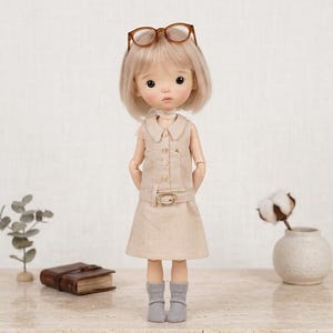 May include: A ball-jointed doll with short blonde hair and brown glasses resting on its head. The doll wears a sleeveless beige collared dress with a matching belt and small grey socks. The doll has a neutral expression and stands upright.