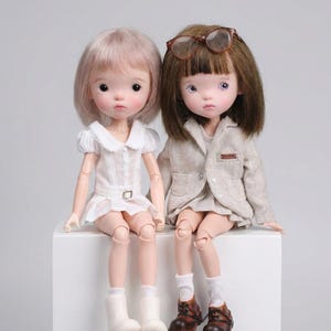 May include: Two ball-jointed dolls with articulated limbs. The left doll has short light pink hair, a white collared dress, and white boots. The right doll has brown hair, brown glasses, a beige blazer over a white top, and brown leather-style shoes.