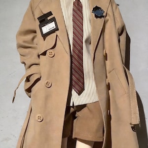 Könnte beinhalten: Ein beigefarbener Trenchcoat mit doppelreihigem Design, getragen über einem gestreiften Hemd und einer bordeauxrot-braun gestreiften Krawatte. Das Outfit umfasst braune Shorts und ein kleines schwarzes Abzeichen. Der Mantel hat ein Revers und Knöpfe.
