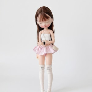 Può includere: Una bambola con lunghi capelli castani indossa un top corto bianco con il logo Adidas, una gonna rosa chiaro a balze e calze bianche al ginocchio con il logo Miu Miu. La bambola ha un orologio nero al polso e una piccola borsa a tracolla beige.