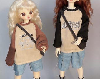 May include: Two dolls wearing casual outfits. One doll has blonde hair and a brown and tan raglan shirt with the word "FRIEND" printed on it. The other doll has red hair and a black and tan raglan shirt with the word "ANTISOCIAL" printed on it. Both dolls wear blue shorts, striped socks, and sneakers.
