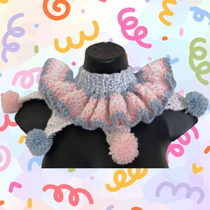 May include: A crocheted ruffled collar featuring a pastel color palette of light blue, soft pink, and white. The accessory includes a ribbed neckband, a wide ruffled edge, and dangling yarn pom-poms in matching blue and pink hues at the ends.