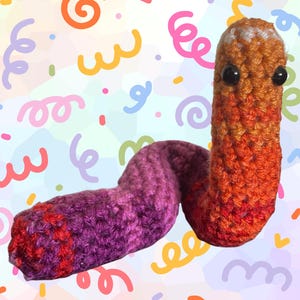 Pode incluir: Um brinquedo de verme de crochê feito à mão em tons de laranja, vermelho e roxo. O verme tem olhos pretos de botão e um corpo curvo. O fundo é um padrão colorido de redemoinhos e confetes.