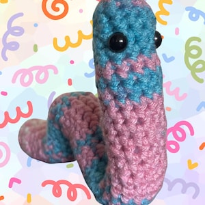 Pode incluir: Um brinquedo de verme de crochê feito à mão em tons de azul e rosa. O verme tem olhos de segurança pretos e é feito com um ponto texturizado. O fundo é um padrão colorido com redemoinhos e confetes.