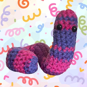 Pode incluir: Um brinquedo de verme de crochê feito à mão em tons de roxo e rosa com olhos de segurança pretos. O verme de pelúcia é enrolado e tem uma aparência texturizada. O fundo apresenta um padrão colorido de confetes.