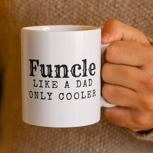 May include: White ceramic mug with the text "Funcle LIKE A DAD ONLY COOLER" in black, held by a person. The mug has a standard handle and a cylindrical shape. The text is in a bold, decorative font.