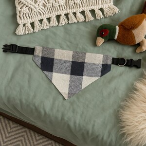 May include: A dog bandana with a black and white checkered pattern, attached to a black adjustable collar with a plastic buckle. A plush toy duck and a macrame pillow are in the background.