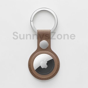 Peut inclure: Un porte-AirTag en cuir marron avec un porte-clés argenté. L'AirTag est argenté et noir avec le logo Apple. Le support a une fermeture à bouton-pression argentée. Le texte "Sunny's Zone" est en filigrane sur l'image.