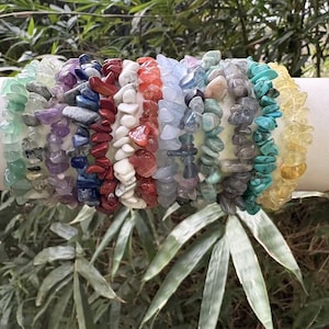 May include: A collection of gemstone chip bracelets in various colors, including green, purple, red, white, orange, blue, gray, turquoise, and yellow. The bracelets are displayed on a white cylinder with gold trim, set against a backdrop of green foliage.