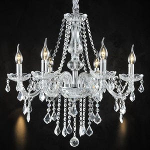 May include: A clear crystal chandelier with six candle-style lights. The fixture has multiple tiers of curved arms and is adorned with hanging crystal droplets and strands. The chandelier is suspended against a black background.