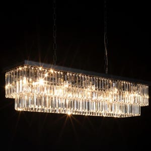 May include: A rectangular chandelier with multiple tiers of clear crystal prisms. The light fixture is suspended from chains and has a dark metal frame. The crystals reflect light, creating a sparkling effect against the black background.