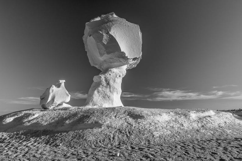 May include: Black and white photograph of a unique rock formation in a desert landscape. The main feature is a large, mushroom-shaped rock balanced on a smaller base. Another smaller rock formation is to the left.