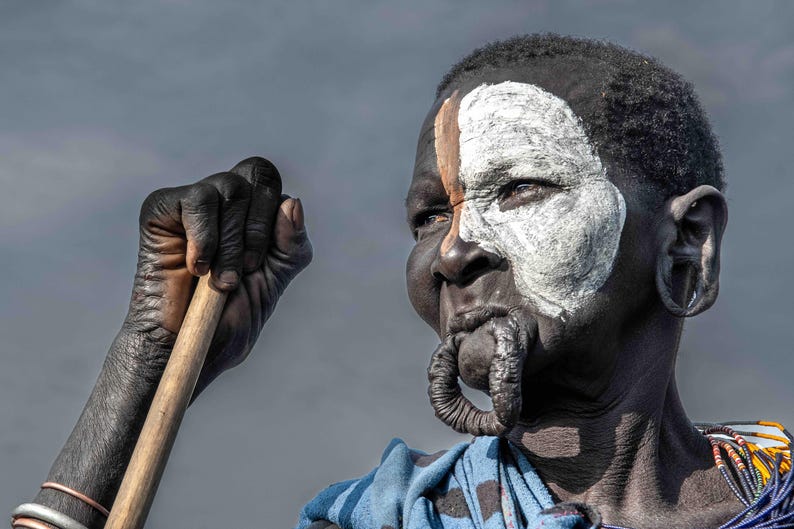 May include: A person with dark skin, adorned with white and brown face paint, and a lip plate. They hold a wooden stick, wearing a blue patterned garment. The person has large ear piercings.
