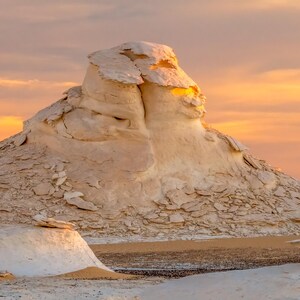 May include: A scenic landscape featuring white rock formations under a warm, orange and yellow sunset sky. The textured rock formations are in the foreground, with a smooth, sandy area in front.