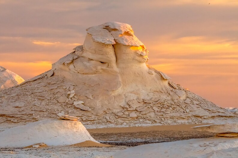 May include: A scenic landscape featuring white rock formations under a warm, orange and yellow sunset sky. The textured rock formations are in the foreground, with a smooth, sandy area in front.