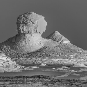 May include: Black and white landscape photograph of a desert scene featuring large, eroded rock formations. The formations have a textured, layered appearance, with a smooth, light-colored surface. The sky is a solid, dark gray.