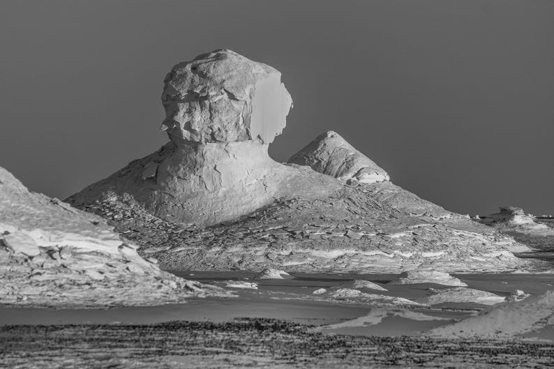 May include: Black and white landscape photograph of a desert scene featuring large, eroded rock formations. The formations have a textured, layered appearance, with a smooth, light-colored surface. The sky is a solid, dark gray.