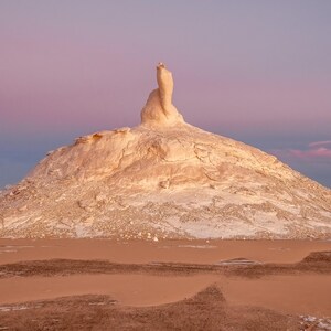 May include: A landscape photograph of a desert scene featuring a large, white rock formation under a pink and blue sky. The foreground shows reddish-brown sand, with the rock formation in the center. The sky transitions from pink to blue.