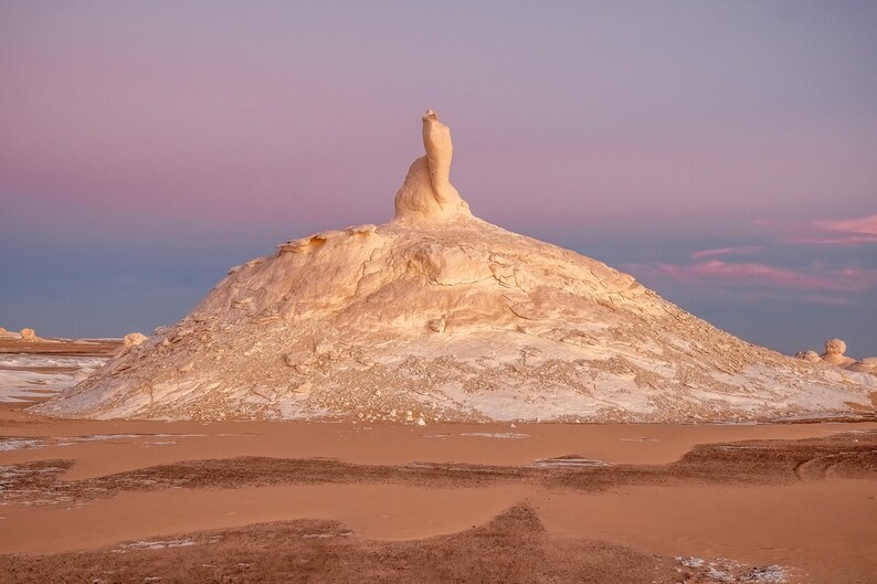 May include: A landscape photograph of a desert scene featuring a large, white rock formation under a pink and blue sky. The foreground shows reddish-brown sand, with the rock formation in the center. The sky transitions from pink to blue.