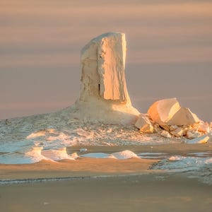 May include: A landscape photograph of a desert scene at sunset. The image features a large, white rock formation in the center, with smaller rock formations and sand in the foreground. The sky is a soft pink and orange.