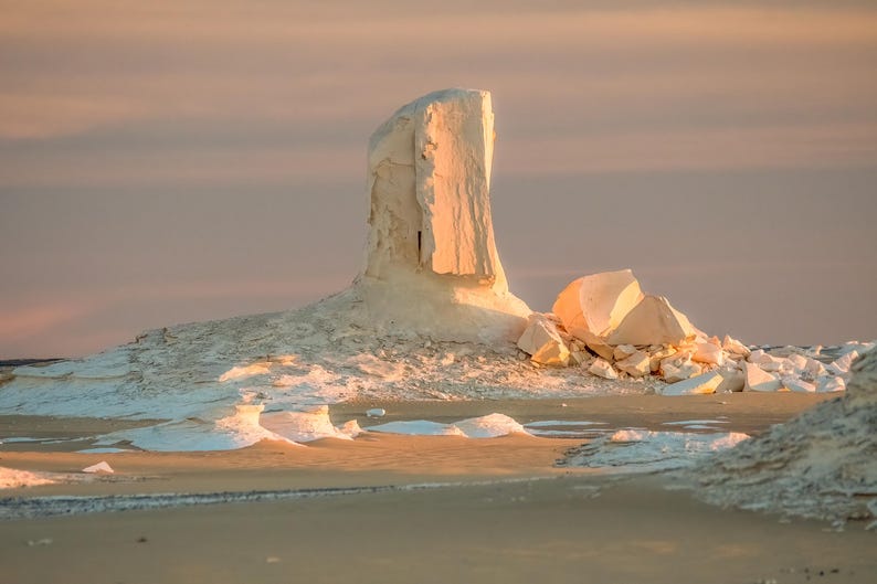 May include: A landscape photograph of a desert scene at sunset. The image features a large, white rock formation in the center, with smaller rock formations and sand in the foreground. The sky is a soft pink and orange.