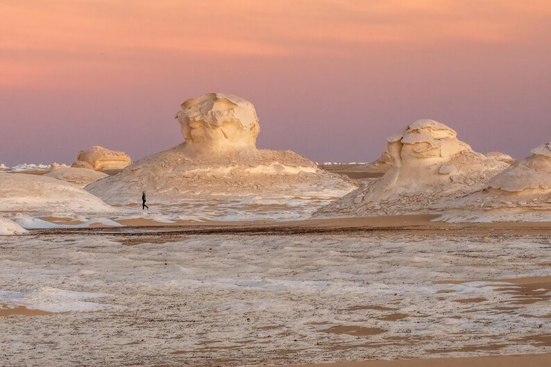 May include: A vast desert landscape under a dusky pink and orange sky. White rock formations dot the sandy terrain, with a lone figure walking in the distance. The scene evokes a sense of solitude and natural beauty.