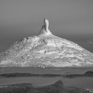 May include: Black and white landscape photograph of a large, white rock formation resembling a mountain. The formation rises from a body of water, with a dark, cloudy sky in the background. The image is a study in contrasts.