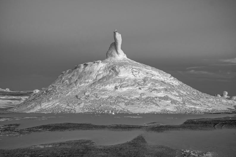May include: Black and white landscape photograph of a large, white rock formation resembling a mountain. The formation rises from a body of water, with a dark, cloudy sky in the background. The image is a study in contrasts.