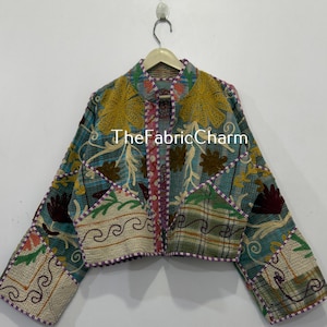Op de afbeelding: Een kort jasje met een patchworkontwerp met diverse bloem- en geometrische patronen in tinten blauw, geel, groen en wit. Het kledingstuk heeft lange mouwen, een opstaande kraag en een knoopsluiting aan de voorzijde met de tekst TheFabricCharm.