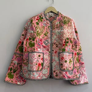 May include: A quilted jacket in a light pink base colour featuring a multi-coloured floral and botanical print. The garment has a round neckline, long sleeves, two front pockets, and a black and white striped trim along the edges and cuffs.