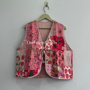 Puede incluir: Un chaleco acolchado sin mangas con un diseño de patchwork en varios tonos de rosa, rojo y blanco con patrones florales y de fresas. El chaleco tiene un cierre de lazo frontal, cuello en V y ribete a rayas blancas y rojas. El texto dice TheFabricCharm.
