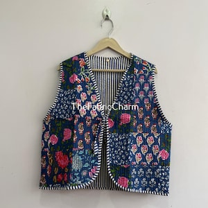 Könnte beinhalten: Eine ärmellose, gesteppte Weste mit einem Patchwork-Design aus blauem Stoff mit rosa und weißen Blumenmustern. Die Weste hat einen V-Ausschnitt, einen Bindegürtel vorne und einen marineblau-weiß gestreiften Saum. Der Text TheFabricCharm ist sichtbar.