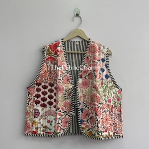 Könnte beinhalten: Eine ärmellose, gesteppte Weste mit einem Patchwork-Blumenmuster in Rot-, Rosa-, Grün- und Orangetönen auf weißem Grund. Die Weste hat einen schwarz-weiß gestreiften Saum und ein Innenfutter sowie einen Bindegürtel auf der Vorderseite. Der Text ThefabricCharm ist sichtbar.