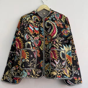 May include: A black quilted jacket featuring a vibrant floral and paisley pattern in shades of red, yellow, blue, and white. The jacket has long sleeves, two front pockets, and a black and white striped trim along the edges and collar.
