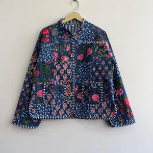 以下が含まれることがあります： ピンク、緑、白の色合いで鮮やかな花柄が施された、濃い青色のキルティングジャケット。マンダリンカラー、長袖、2つのフロントパッチポケットが特徴で、縁には青と白のストライプのトリミングが施されています。