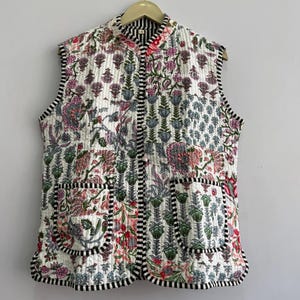 Puede incluir: Un chaleco acolchado sin mangas con base blanca y un estampado floral multicolor en tonos rosa, verde y azul. La prenda incluye dos bolsillos delanteros de parche y está rematada con un ribete a rayas blancas y negras en los bordes.