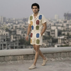 May include: A cream-colored short-sleeved shirt and shorts set. The outfit features colorful, rectangular patches in blue, red, green, and yellow. The person is wearing beige sneakers. The background shows a cityscape.