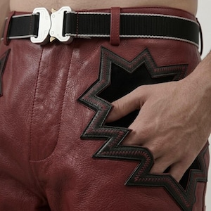 Puede incluir: Primer plano de pantalones de cuero rojos con detalles en forma de estrella negra y un cinturón a rayas blanco y negro. El cinturón tiene una hebilla plateada. Una mano está en el bolsillo.