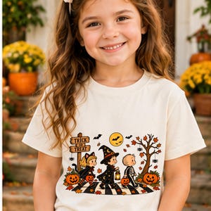 May include: A cream-coloured t-shirt featuring a graphic illustration of three children in Halloween costumes. The design includes a wooden sign reading Trick or Treat, a moon, bats, a tree, pumpkins, and autumn leaves on a striped path.