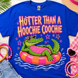 Op de afbeelding: Koningsblauw T-shirt met de tekst "Hotter Than A Hoochie Coochie" in geel en roze. Het shirt heeft een cartoon alligator met een zonnebril die op een zwemband drijft. Roze en oranje armbanden en zonnebrillen zijn zichtbaar.
