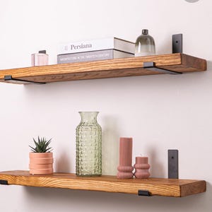 May include: Two wooden floating shelves with black metal brackets are mounted on a white wall. The top shelf holds books and a glass cloche. The bottom shelf displays a potted succulent, a textured glass vase, and decorative candles.