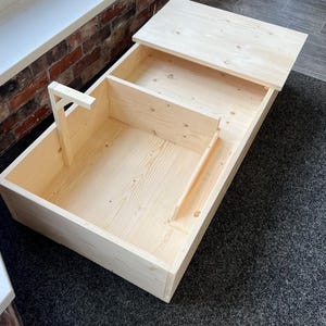 Könnte beinhalten: Eine unfertige Holzkiste mit hellem Holz, rechteckig, mit aufklappbarem Deckel und Innenteilern. Die Kiste ist aus hellem Holz, möglicherweise Kiefer, gefertigt. Der Deckel ist teilweise geöffnet und zeigt die Innenfächer. Für die Aufbewahrung konzipiert.