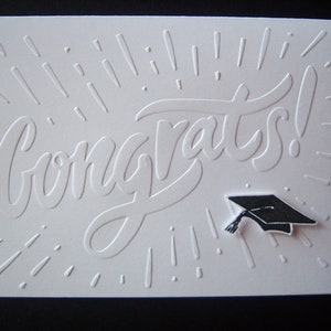 May include: White card with the word "Congrats!" embossed in a stylized font. A black graduation cap is embossed in the lower right corner.