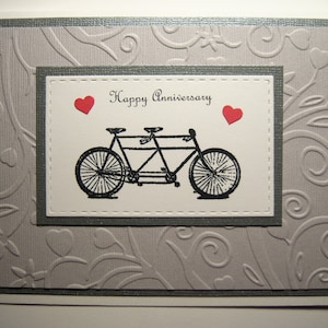 May include: A gray card with a white inner border and a gray inner border. The inner border has a white background with a black and white illustration of a tandem bicycle. The text "Happy Anniversary" is printed in black above the bicycle. Two red hearts are on either side of the text.